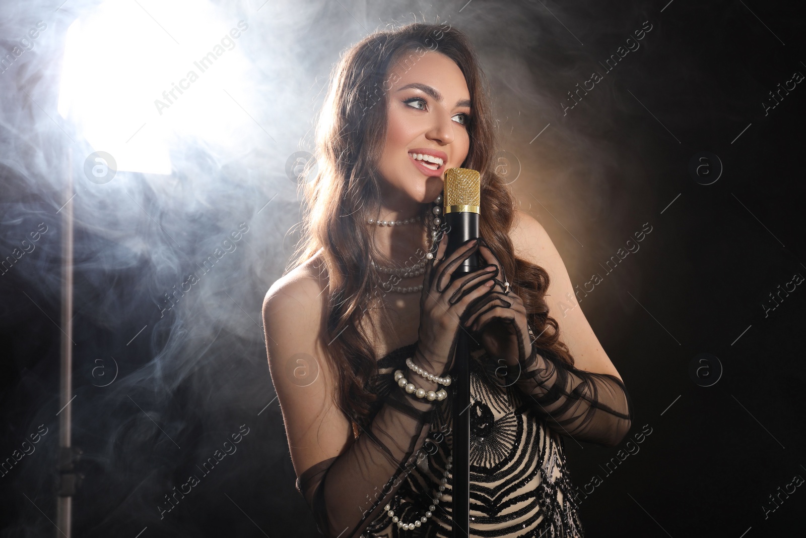 Photo of Beautiful young woman with microphone singing on dark background with smoke