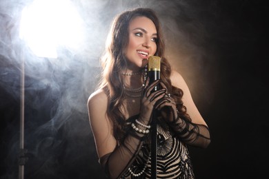 Beautiful young woman with microphone singing on dark background with smoke