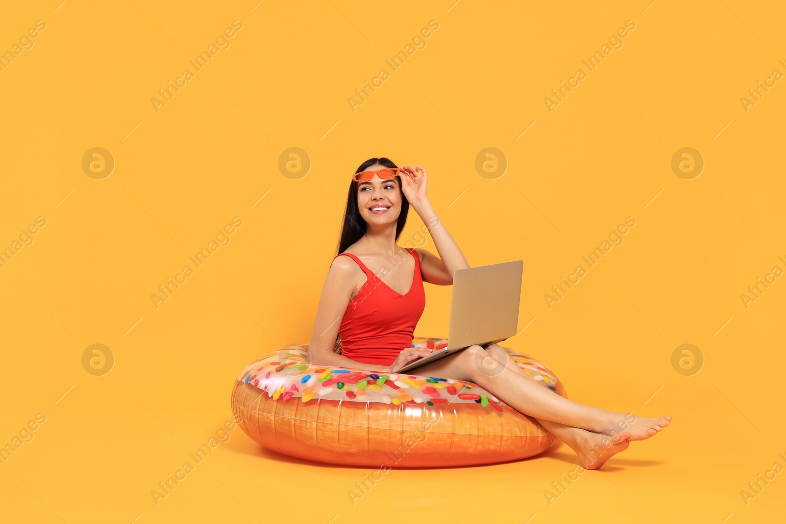 Photo of Happy young woman with beautiful suntan using laptop on inflatable ring against orange background