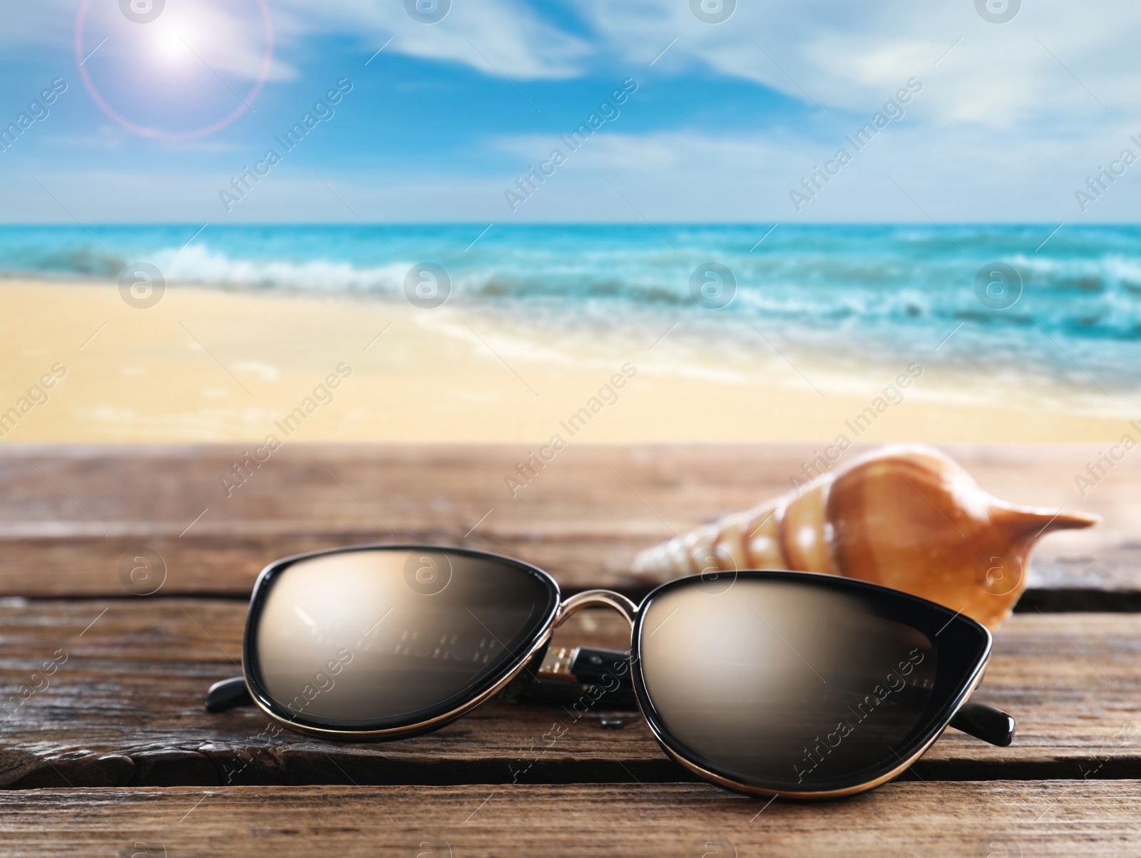 Image of Shell and stylish sunglasses on wooden table near sea with sandy beach