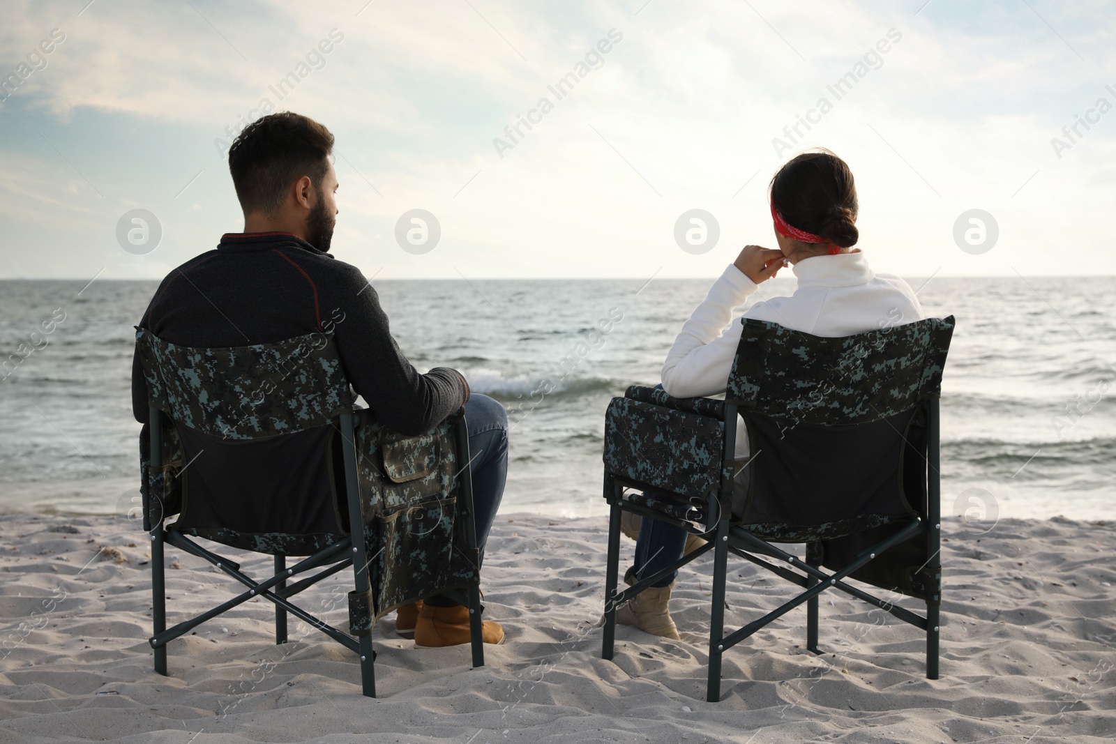 Photo of Couple sitting in camping chairs and enjoying seascape on beach, back view