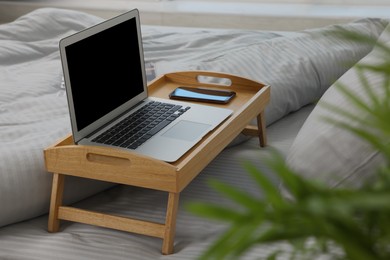 Photo of Wooden tray table with laptop and smartphone on bed indoors