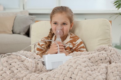 Photo of Little girl using nebulizer for inhalation in armchair at home