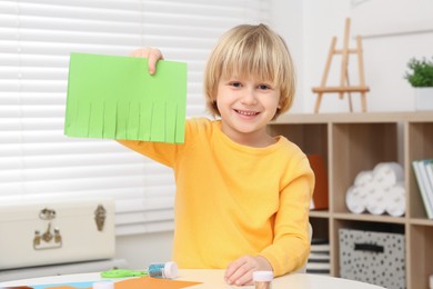Cute little boy with cut green paper at desk in room. Home workplace