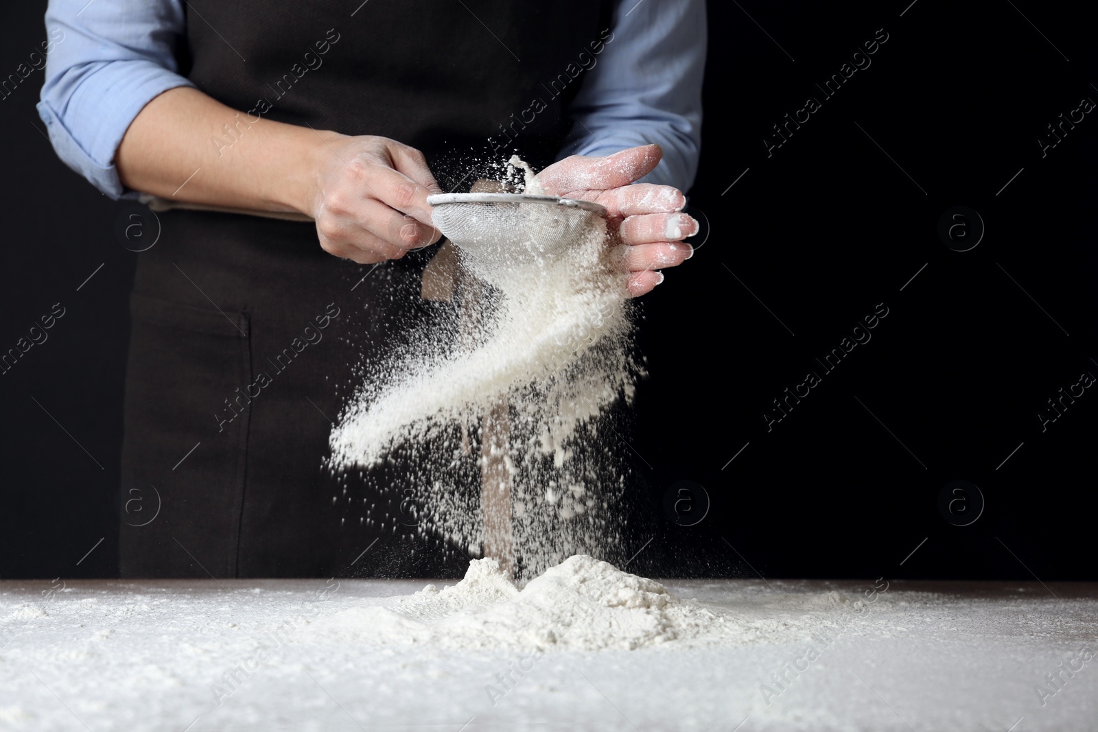 Photo of Woman sifting wheat flour at table against black background, closeup