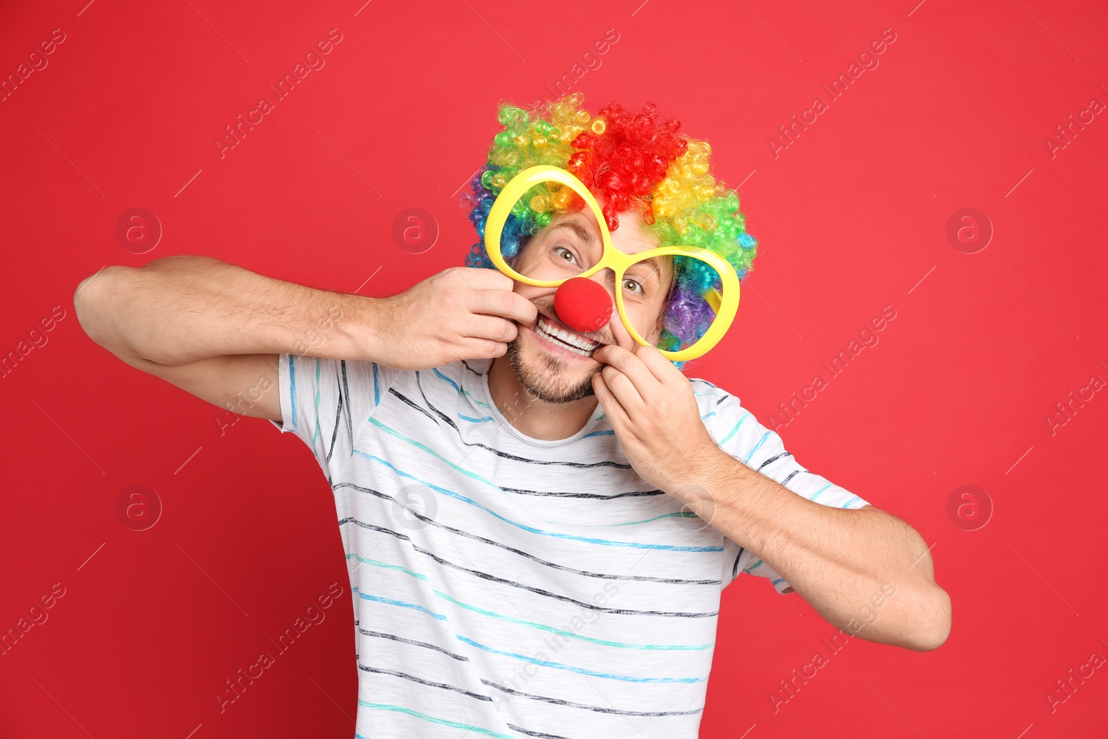 Photo of Funny man with large glasses, rainbow wig and clown nose on red background. April fool's day