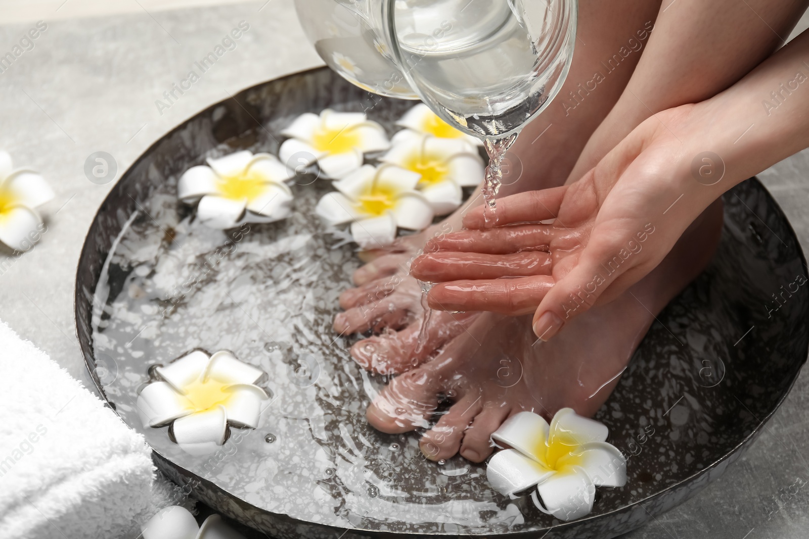 Photo of Woman pouring water onto hand while soaking her feet in bowl on light grey floor, closeup. Spa treatment