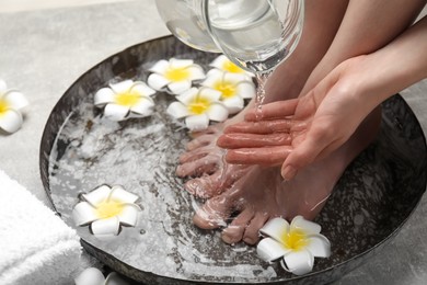 Photo of Woman pouring water onto hand while soaking her feet in bowl on light grey floor, closeup. Spa treatment