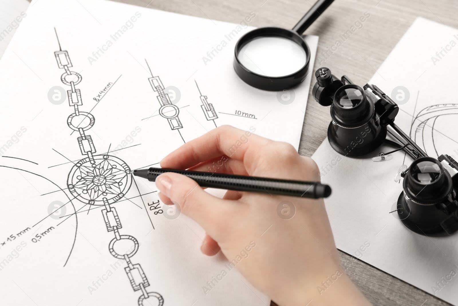 Photo of Jeweler drawing sketch of elegant bracelet on paper at wooden table, closeup