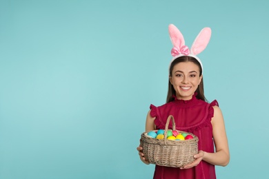 Photo of Beautiful woman in bunny ears headband holding basket with Easter eggs on color background, space for text