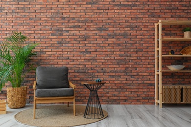 Modern room interior with stylish grey armchair and potted plant near brick wall