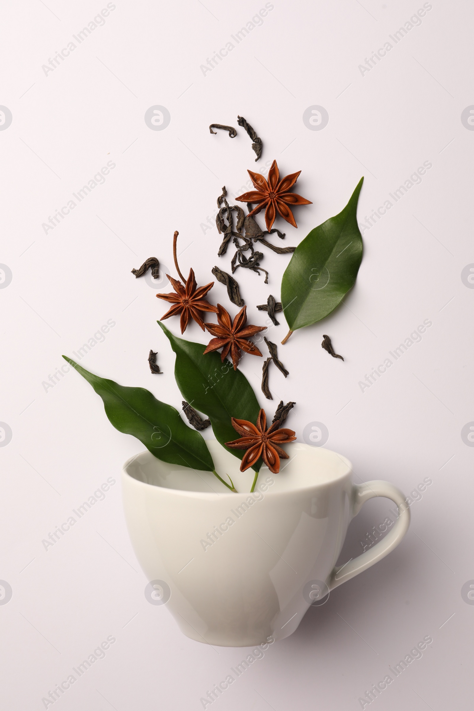 Photo of Anise stars, dry tea and green leaves falling into cup on white background, flat lay