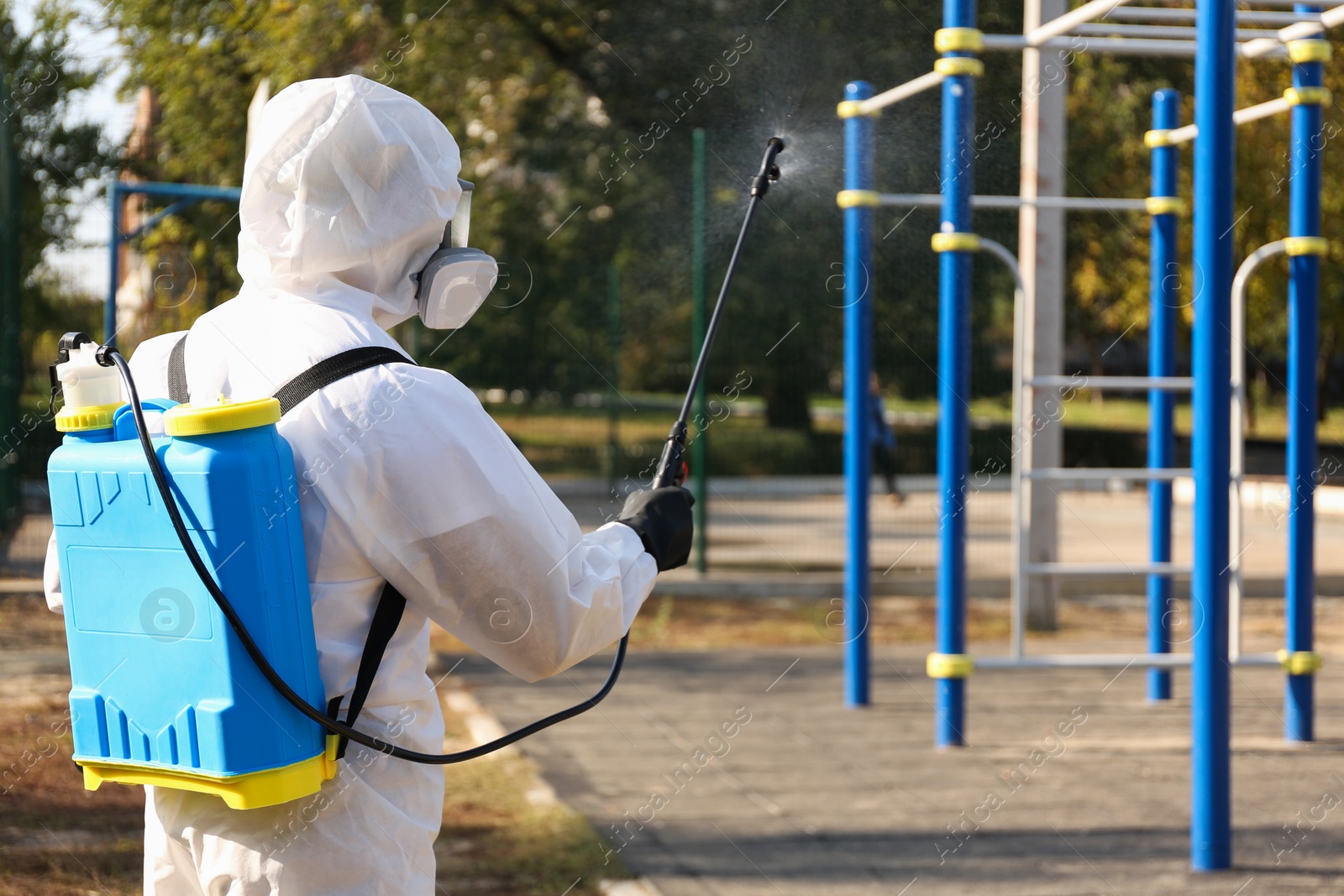 Photo of Person in hazmat suit spraying disinfectant around outdoor gym. Surface treatment during coronavirus pandemic