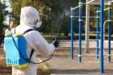 Photo of Person in hazmat suit spraying disinfectant around outdoor gym. Surface treatment during coronavirus pandemic