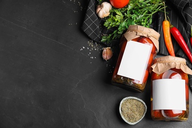 Photo of Jars of pickled tomatoes with blank stickers on black table, flat lay. Space for text