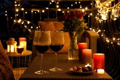 Photo of Vase with roses, glasses of wine, snacks and candles on rattan table at balcony in night