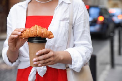 Photo of Woman holding tasty croissant and cup of coffee on city street, closeup