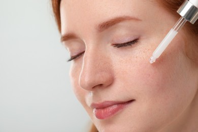 Photo of Beautiful woman with freckles applying cosmetic serum onto her face against grey background, closeup