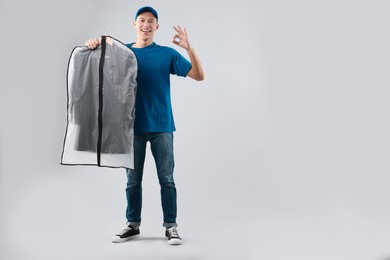 Photo of Dry-cleaning delivery. Happy courier holding garment cover with clothes and showing OK gesture on light grey background, space for text
