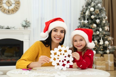 Photo of Mother and daughter in Santa hats with paper snowflake near Christmas tree at home