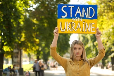 Photo of Sad woman holding poster in colors of national flag and words Stand with Ukraine outdoors. Space for text