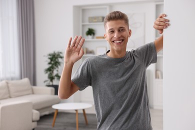 Photo of Happy man waving near white wall at home, space for text. Invitation to come in room