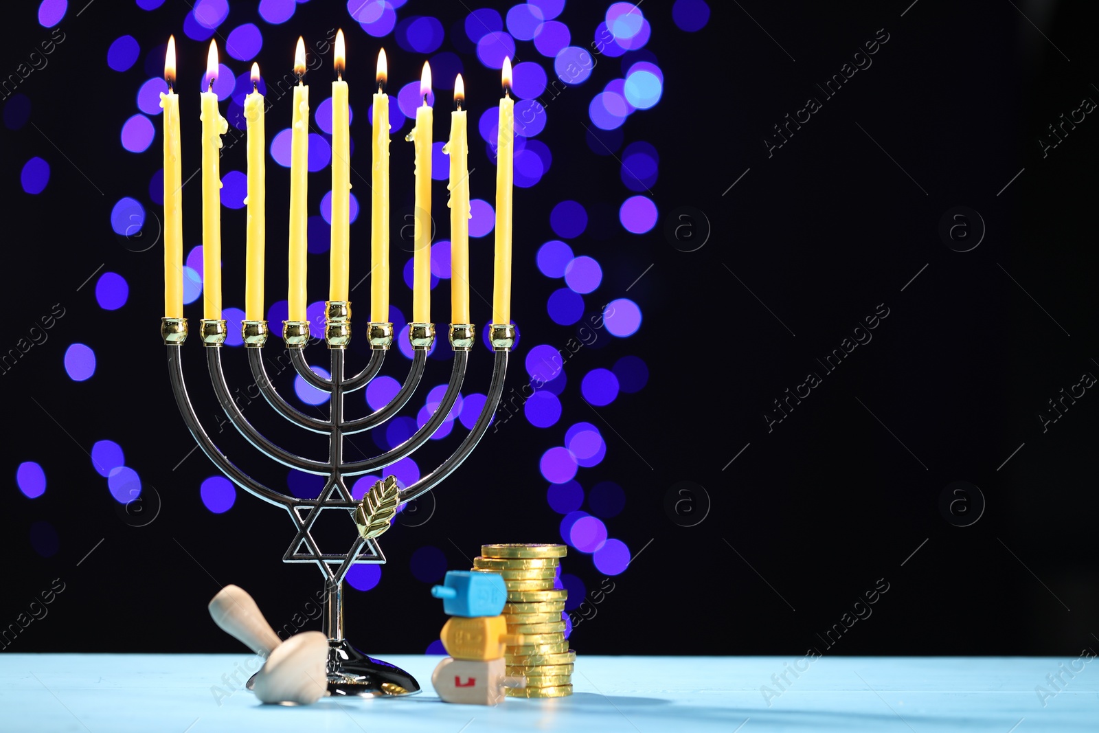 Photo of Hanukkah celebration. Menorah with burning candles, dreidels and golden coins on light blue table against blurred lights, space for text