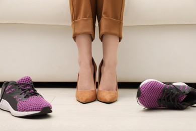 Photo of Woman in high heel shoes sitting on sofa near comfortable sneakers in office, closeup