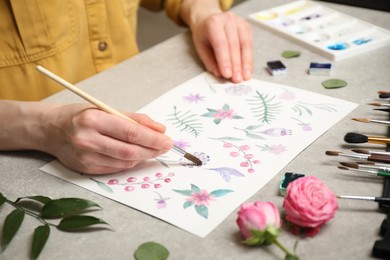 Photo of Woman painting flowers with watercolor at grey stone table, closeup
