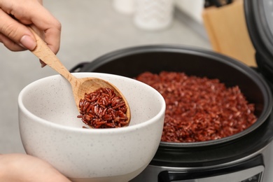 Photo of Woman putting brown rice into bowl from multi cooker in kitchen, closeup