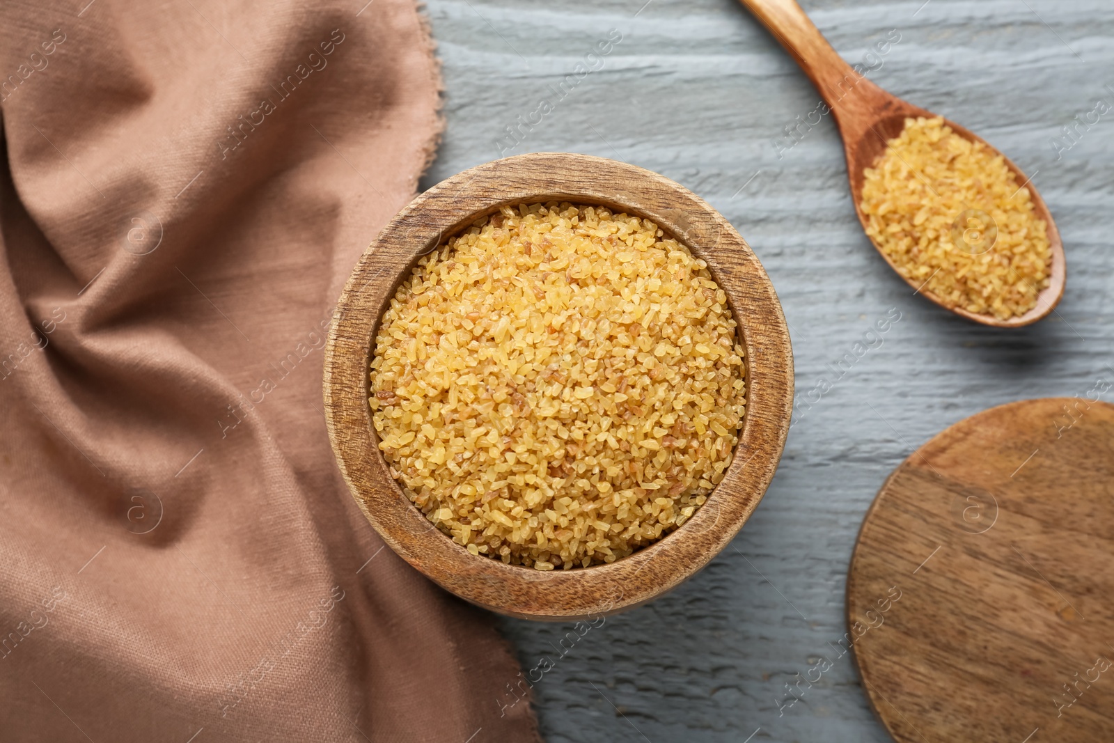 Photo of Bowl and spoon with uncooked bulgur on grey wooden table, flat lay