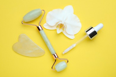 Photo of Gua sha stone, face roller, dropper and orchid flower on yellow background, flat lay