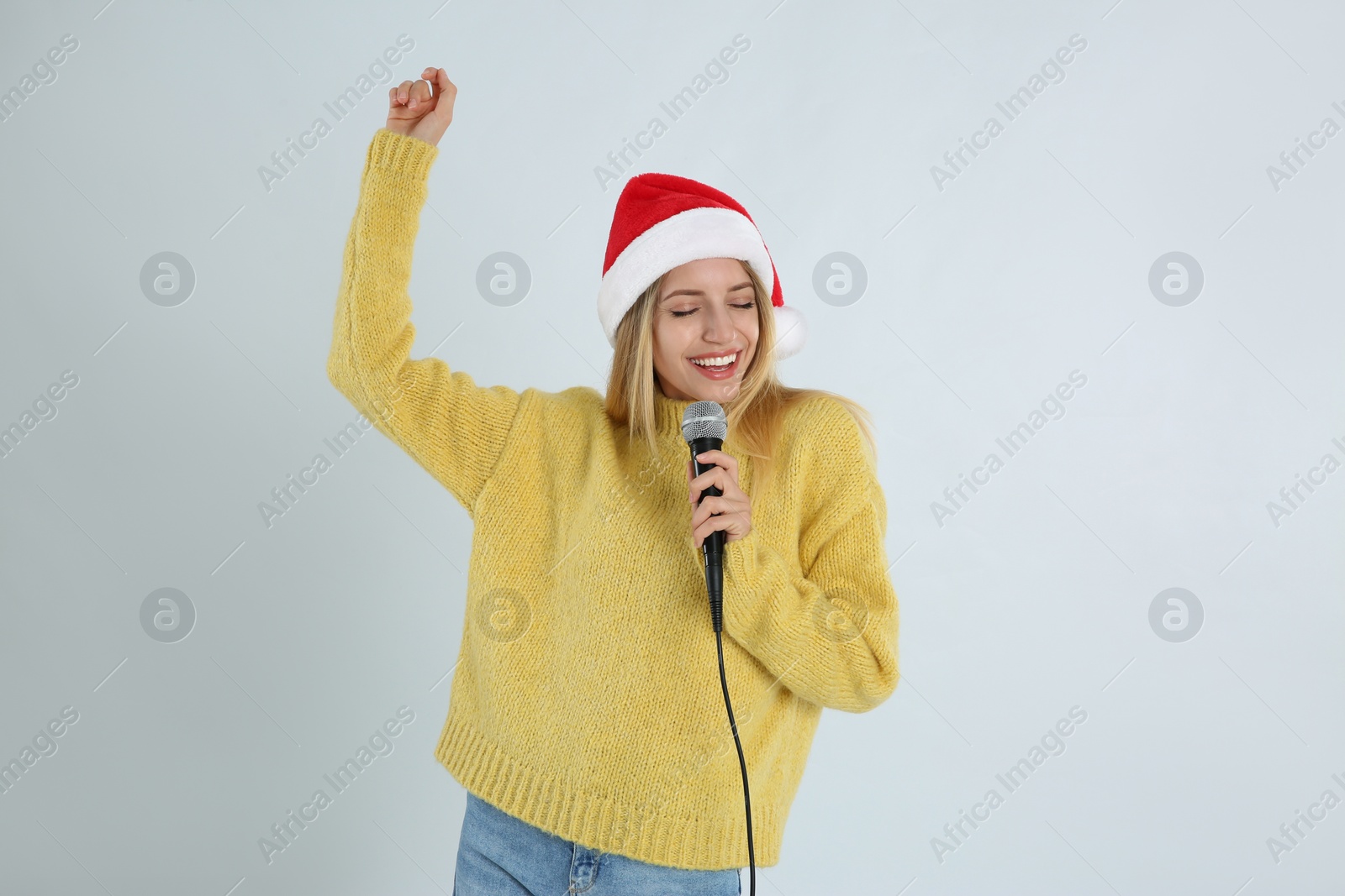 Photo of Emotional woman in Santa Claus hat singing with microphone on light grey background. Christmas music