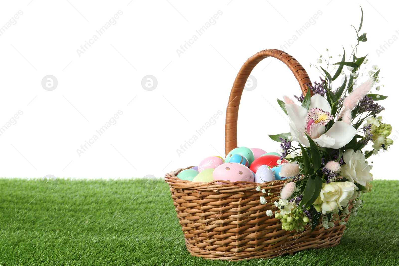 Photo of Wicker basket with Easter eggs and beautiful flowers on green grass against white background. Space for text