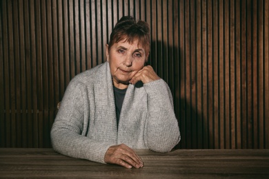 Portrait of poor senior woman sitting at table against wooden wall