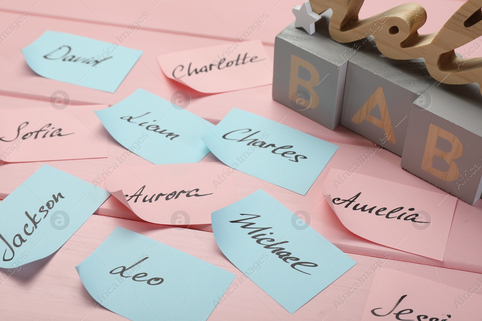 Photo of Cubes with word Baby and paper stickers with different names on pink wooden table. Choosing baby's name