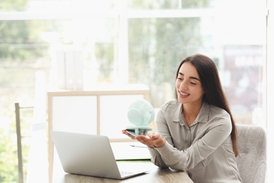 Photo of Young woman enjoying air flow from portable fan at workplace, space for text. Summer heat