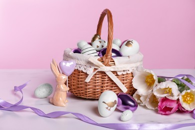 Photo of Wicker basket with festively decorated Easter eggs, bunny and beautiful tulips on white marble table against pink background