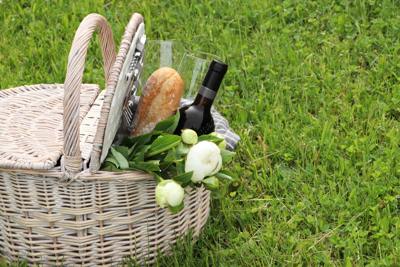 Photo of Picnic basket with wine, bread and flowers on green grass outdoors