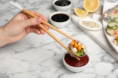 Photo of Woman dipping delicious roll wrapped in rice paper into sauce at white marble table, closeup