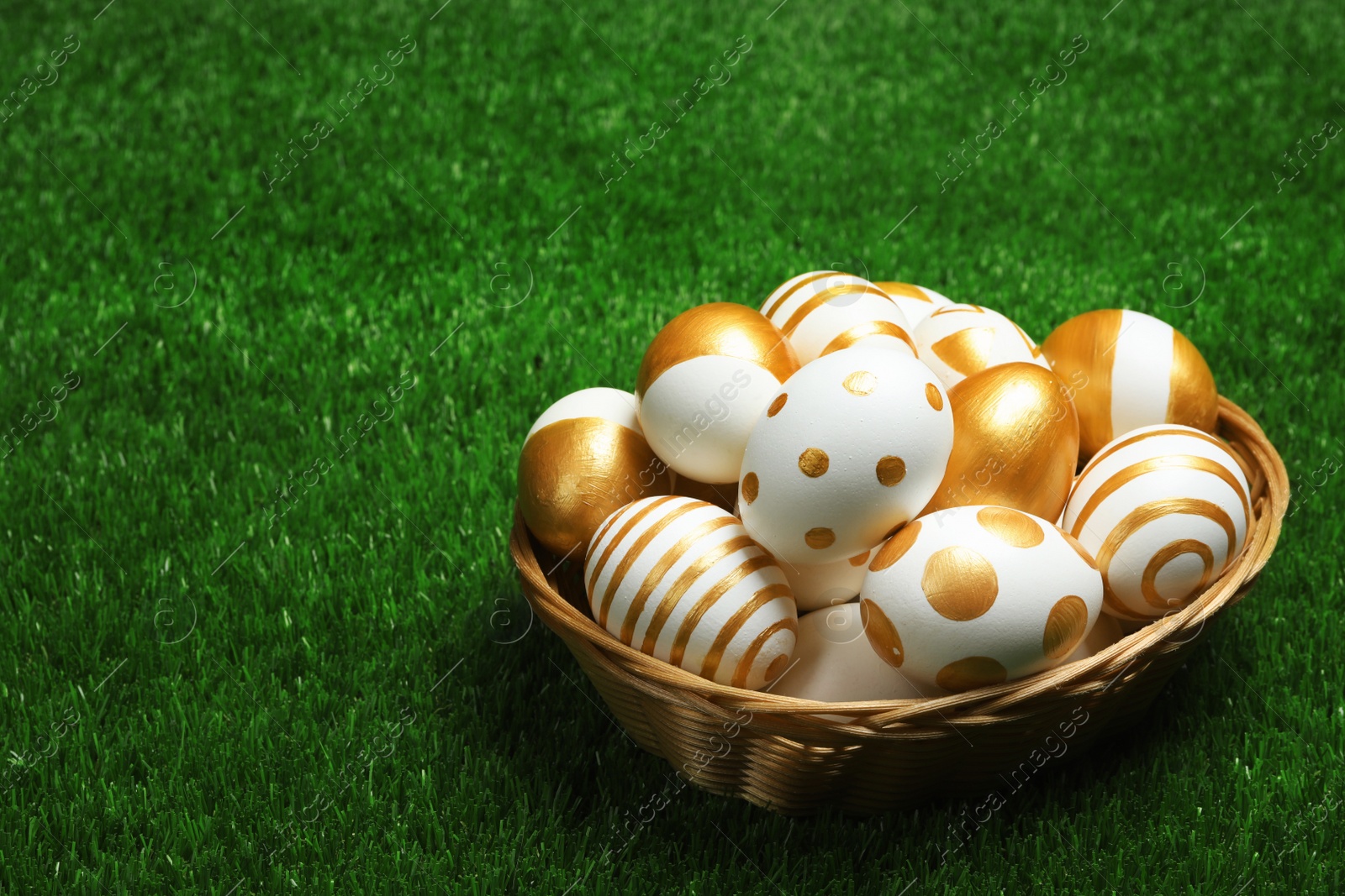 Photo of Wicker basket of traditional Easter eggs decorated with golden paint on green lawn, space for text