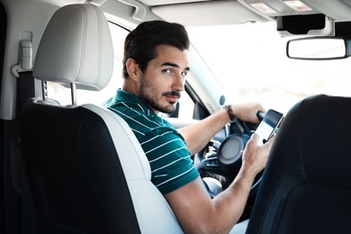 Photo of Attractive young man with smartphone driving luxury car, view from backseat