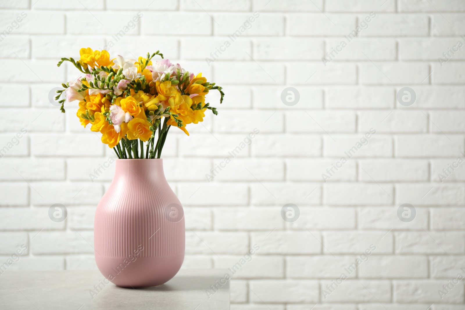 Photo of Beautiful blooming freesias in pink vase on table against white brick wall. Space for text