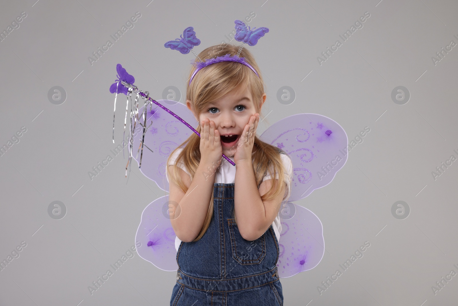 Photo of Surprised little girl in fairy costume with violet wings and magic wand on light grey background