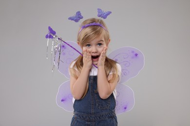 Surprised little girl in fairy costume with violet wings and magic wand on light grey background