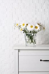Photo of Beautiful tender chamomile flowers in vase on wooden commode near white textured wall