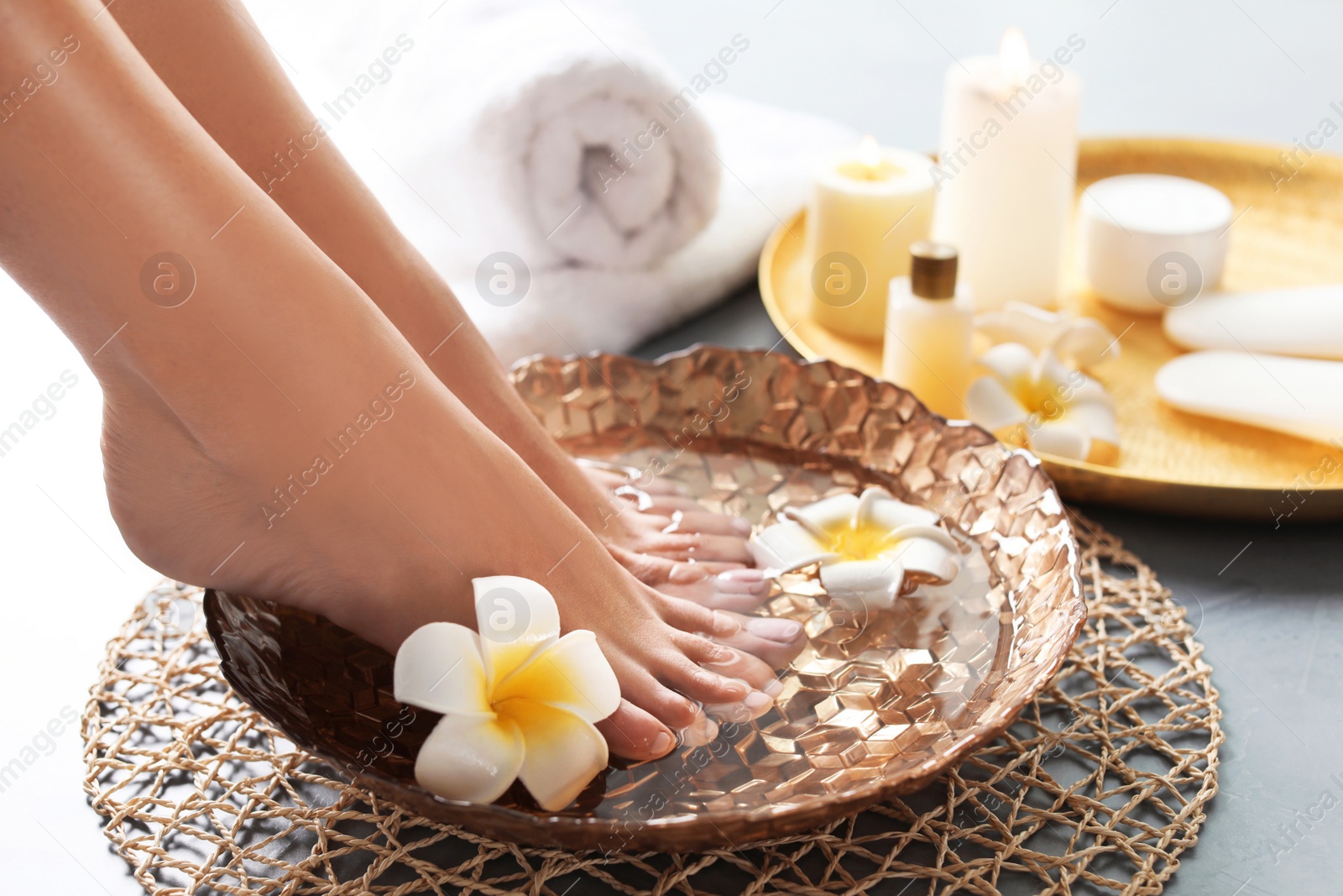 Photo of Closeup view of woman soaking her feet in dish with water and flowers on grey floor. Spa treatment