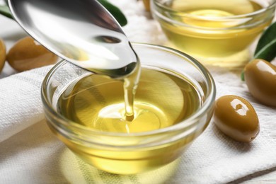 Photo of Spoon with cooking oil over bowl and olives on white wooden table, closeup