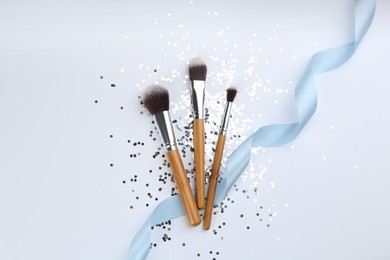 Photo of Different makeup brushes, ribbon and shiny confetti on white background, flat lay