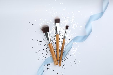 Different makeup brushes, ribbon and shiny confetti on white background, flat lay